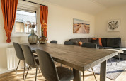 Lichte lodge woonkamer met houten tafel, zwarte stoelen, oranje gordijnen en moderne muurdecoratie.