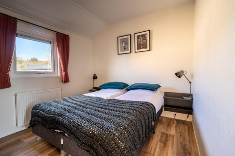 Dormitorio luminoso en una cabaña con cama doble, mesillas, lámparas, ventana y suelo de madera.