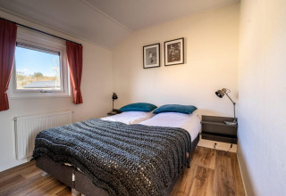 Dormitorio luminoso en una cabaña con cama doble, mesillas, lámparas, ventana y suelo de madera.