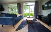 Moderne stue i Just Nature Lodge, Limburg, Holland, med blå sofa, store vinduer og smukt udsyn.