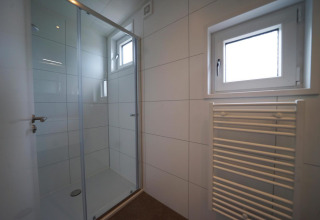 Moderne douche met glazen deur, witte tegels en radiator bij venster in Just nature lodge Limburg.