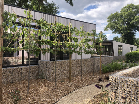 Moderne lodge ved Just Nature i Limburg, Holland, omgivet af træer, sten strukturer og havegang.