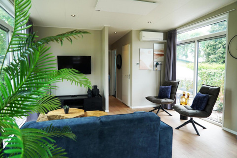 Moderne woonkamer in Just Nature Lodge te Limburg, Nederland, met grote ramen, blauwe zetel en planten.
