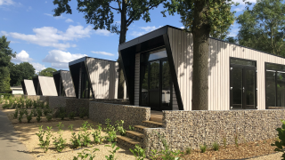 Lodges Just Nature Light modernes avec de grandes fenêtres, murs en pierre et verdure sous un ciel bleu.