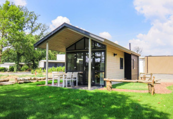 Billede af Tiny House-lodge med overdækket terrasse, havebord og stole på en grøn græsplæne.