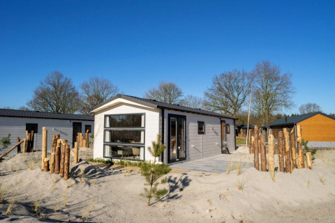 Moderne Berkel 4 Lodge auf sandigem Gelände mit Holzzaun, Bäumen und klarem Himmel im Hintergrund.