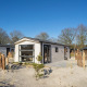 Moderne lodge Berkel 4 på sandet grund med træhegn og træer i baggrunden under blå himmel.
