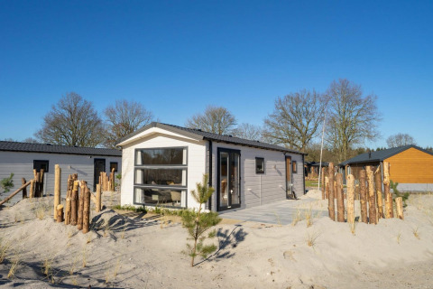 Moderne Berkel 4 Lodge auf sandigem Gelände mit Holzzaun, Bäumen und klarem Himmel im Hintergrund.