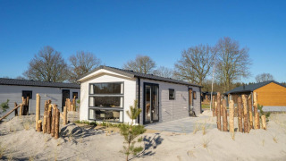 Moderne Berkel 4 Lodge auf sandigem Gelände mit Holzzaun, Bäumen und klarem Himmel im Hintergrund.