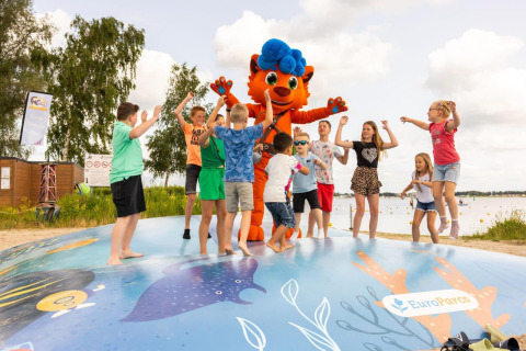 Des enfants jouent et sautent avec une mascotte sur une aire de jeux près d’un lac en glamping.