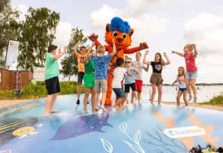 Kinderen spelen en springen met een mascotte op een speelkoepel bij een glamping-accommodatie aan het meer.