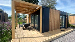 Lodge moderne Domus 4 avec façade en bois, terrasse extérieure, pergola et mobilier de jardin, cadre verdoyant.