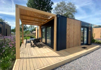 Moderne Lodge Domus 4 mit Holzverkleidung, Terrasse, Pergola und Gartenmöbeln in naturnaher Umgebung.