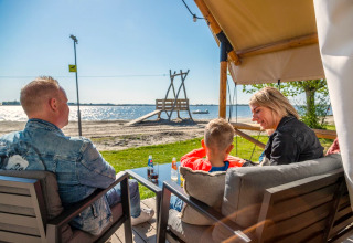 Een gezin ontspant buiten hun glampingtent met uitzicht op het strand en het water bij zonnig weer.