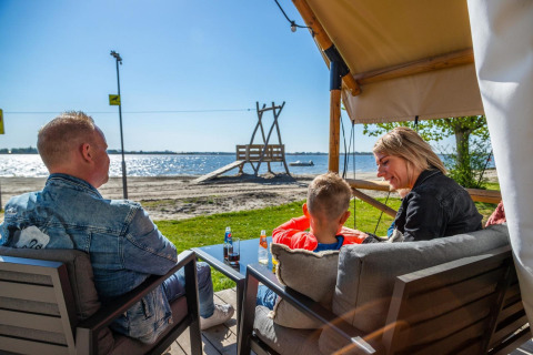 Une famille se détend devant sa tente de glamping avec vue sur la plage et l’eau scintillante.