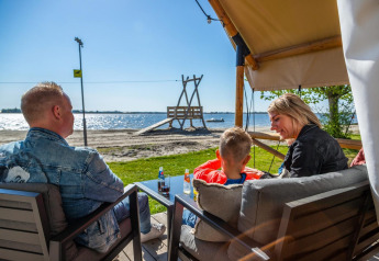 Une famille se détend devant sa tente de glamping avec vue sur la plage et l’eau scintillante.