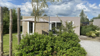 Cosy Unique 2+4 lodge met moderne uitstraling, parasol en veel groen op de voorgrond in een tuinsetting.