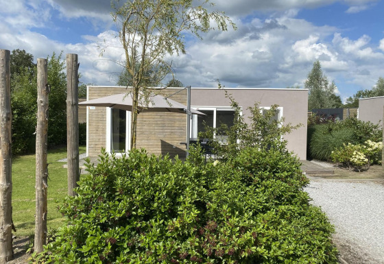 Cosy Unique 2+4 lodge met moderne uitstraling, parasol en veel groen op de voorgrond in een tuinsetting.