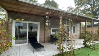Overdekt terras van Cosy Unique 4 lodge met houten vloer, ligzetel, tuinmeubilair en omliggend groen.