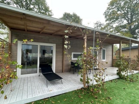 Cosy Unique 4 lodge exterior with covered wooden deck, lounge chair, table and chairs, surrounded by greenery.