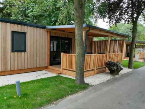 Moderne træhytte, Cottage 5, med overdækket terrasse, grøn græsplæne og træer i naturskønne omgivelser.