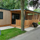 Moderne træhytte, Cottage 5, med overdækket terrasse, grøn græsplæne og træer i naturskønne omgivelser.