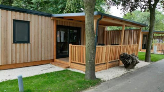 Zeitgemäße Lodge Cottage 5 aus Holz mit überdachter Terrasse, viel Grün und Bäumen in ruhiger Lage.