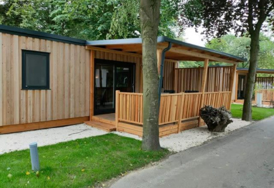 Moderne træhytte, Cottage 5, med overdækket terrasse, grøn græsplæne og træer i naturskønne omgivelser.
