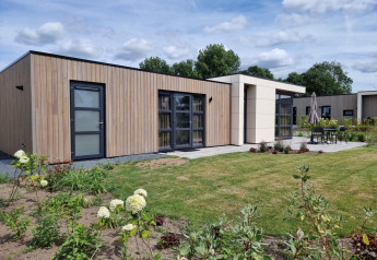 Cube Maximaal 4 Lodge mit moderner Holzfassade, großen Fenstern und gepflegtem Garten vor blauem Himmel.