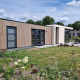Cube Maximaal 4 Lodge mit moderner Holzfassade, großen Fenstern und gepflegtem Garten vor blauem Himmel.