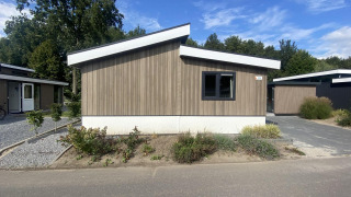 Modernes Hackfort MIVA-Lodge mit Holzverkleidung, schrägem Dach und grüner Bepflanzung an einem sonnigen Tag.