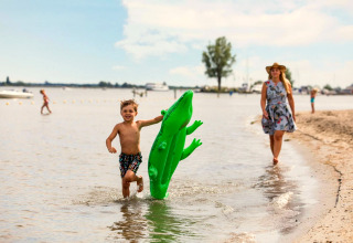Junge spielt mit grüner aufblasbarer Krokodil am Strand, Frau im Kleid und Hut läuft dahinter.