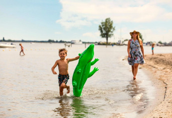 Un garçon court avec un crocodile gonflable vert près de l’eau, suivi par une femme en robe et chapeau.