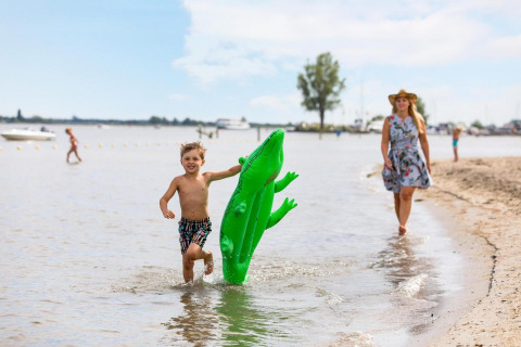 Jongen loopt met groene opblaaskrokodil in het water, vrouw in zomerjurk en hoed volgt op het strand.