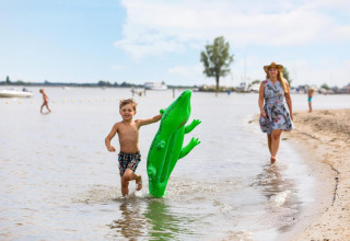 Jongen loopt met groene opblaaskrokodil in het water, vrouw in zomerjurk en hoed volgt op het strand.