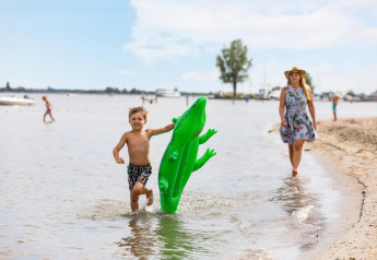 Jongen loopt met groene opblaaskrokodil in het water, vrouw in zomerjurk en hoed volgt op het strand.