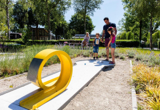 Famiglia che gioca a mini golf all'aperto in un glamping, circondata dal verde e sotto il sole.