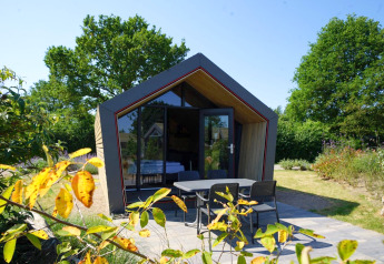 Moderne Solo Retreat 2-lodge med stor glasfacade, hyggelig terrasse og grønne omgivelser i sommersolen.