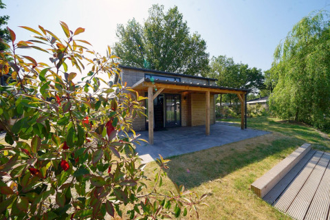 Kleines Tiny House Plus 4 Lodge im Sonnenschein mit Holzterrasse und grünem Garten, umgeben von Bäumen