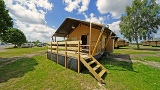 Glampingtent 6 lodge with wooden deck at a campground, surrounded by green fields and other cabins.