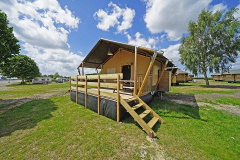 Glampingtent 6 Lodge mit Holzterrasse auf einem Campingplatz, umgeben von Grünflächen und anderen Zelten.