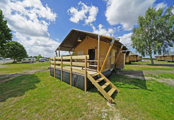 Glampingtent 6 lodge with wooden deck at a campground, surrounded by green fields and other cabins.