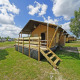 Glampingtent 6 lodge med træterrasse på en campingplads, omgivet af grønne områder og andre telte.