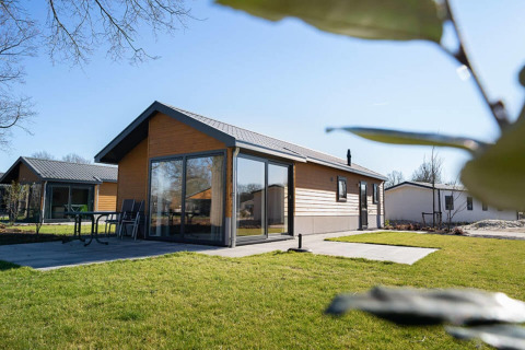 Lodge moderno con grandi finestre e patio, circondato da prato verde e cielo azzurro in una giornata di sole.
