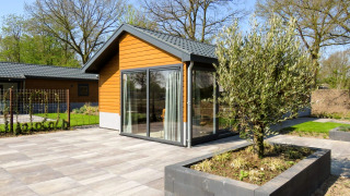 Lodge moderne Velthorst 6 avec de grandes baies vitrées, terrasse et arbre au cœur d’un jardin paysager.