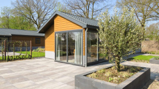 Moderne lodge ved Velthorst 6 med store vinduer, terrasse og træ i en frodig, grøn have om foråret.