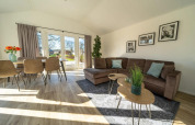 Lyst og moderne lodge-stue med hjørnesofa, spisebord, store vinduer og udsigt til have og terrasse.