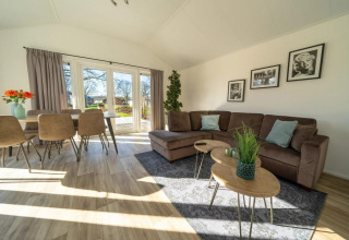 Salon lumineux et moderne de lodge avec canapé d’angle, table à manger, grandes fenêtres et vue jardin.