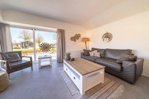Salon de lodge lumineux et moderne avec grandes fenêtres, canapé en cuir, fauteuil et vue sur la nature.
