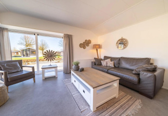 Lichte en moderne woonkamer in lodge met grote ramen, leren zetel, fauteuil en zicht op de natuur.